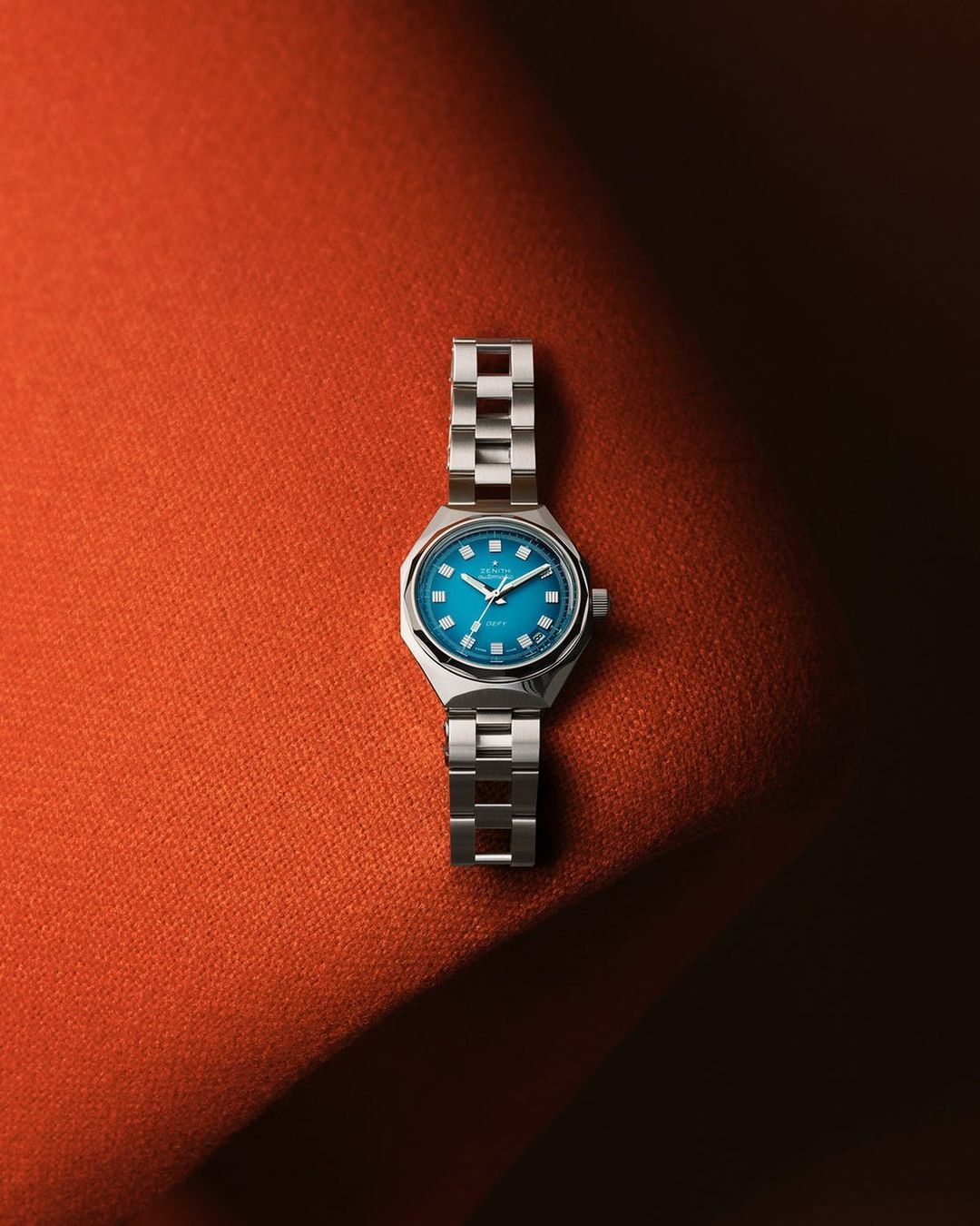 Wristwatch with a silver band and a teal face displayed on a textured orange surface with an eye-level angle and studio lighting.