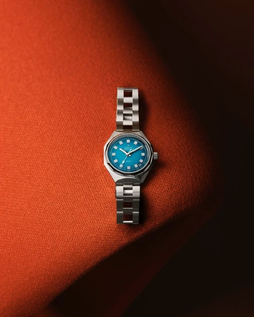 Wristwatch with a silver band and a teal face displayed on a textured orange surface with an eye-level angle and studio lighting.
