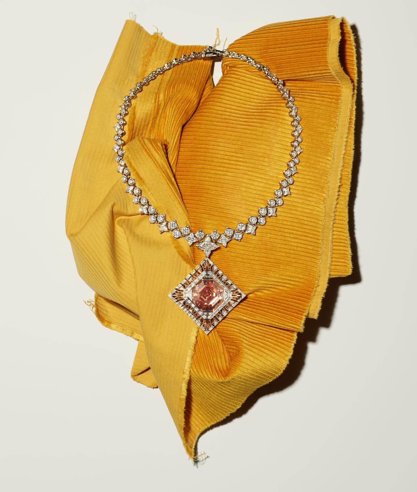 Elegant diamond necklace featuring a large pink diamond pendant, displayed on a mustard-yellow textured cloth against a plain white background.