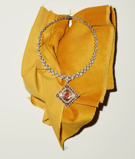 Elegant diamond necklace featuring a large pink diamond pendant, displayed on a mustard-yellow textured cloth against a plain white background.