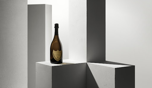 A bottle of Dom Pérignon champagne sits on a geometric white cube in a bright studio setting with vertical columns in the background.