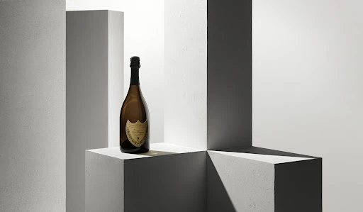 A bottle of Dom Pérignon champagne sits on a geometric white cube in a bright studio setting with vertical columns in the background.