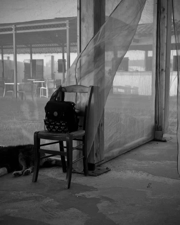 Black and white photograph of a dark wooden chair with a backpack resting on it under a shelter with a billowing sheer curtain.
