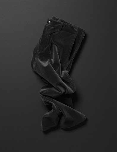 Elevated studio shot of folded, dark corduroy pants on a black background with soft shadows. Minimalist fashion photography.
