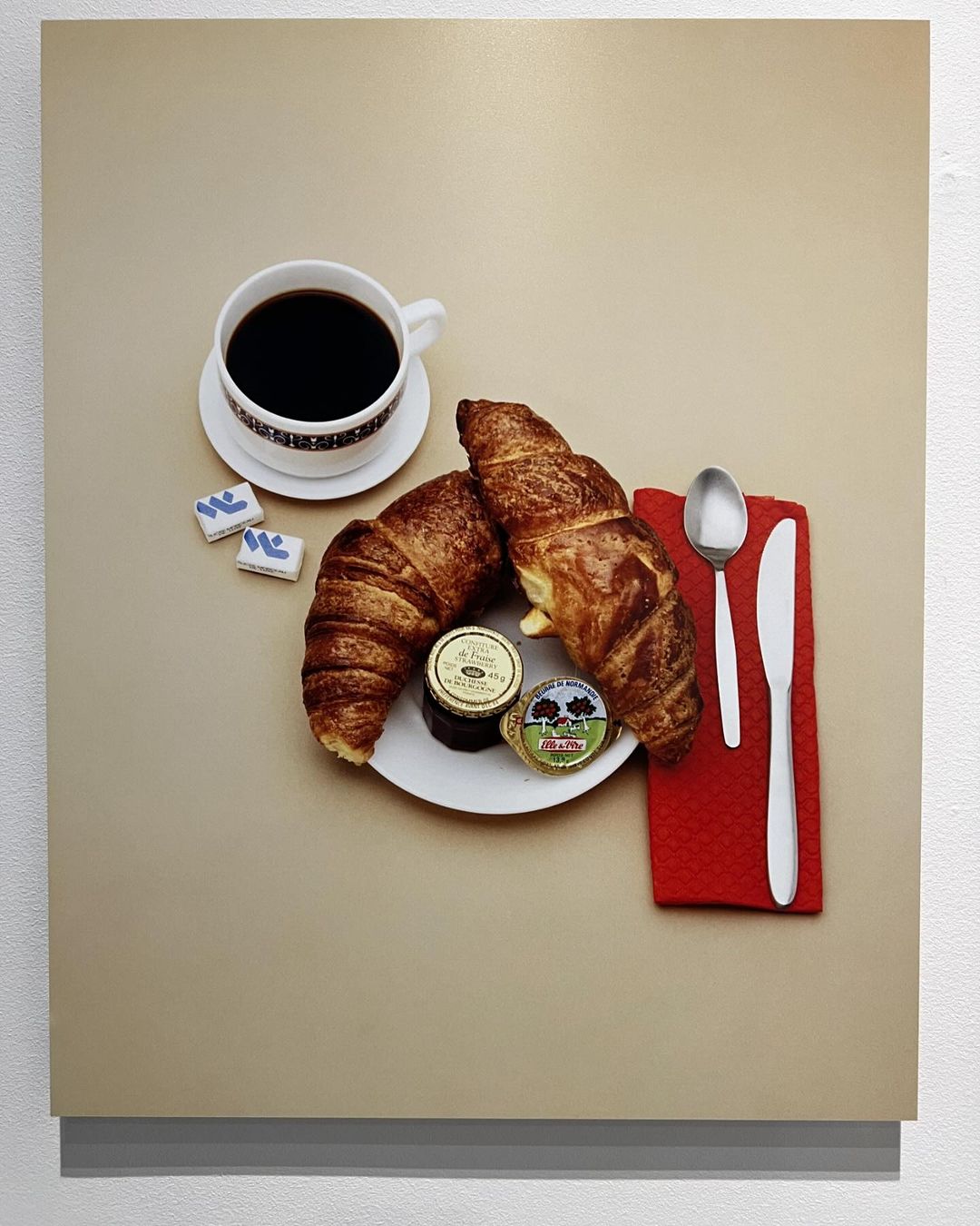 Still life of a coffee cup with dark liquid, pastries, preserves on a white plate, silverware, and colorful tiles on a tan background.