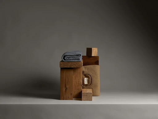 Still life of a folded gray blanket and stacked wooden blocks and a brown textile bag on a light gray surface against a dark gray background.