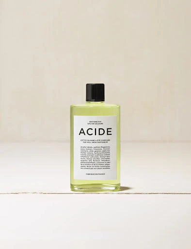 Close-up studio shot of a bottle of ACIDE eau de cologne on a white surface with a neutral-toned background.