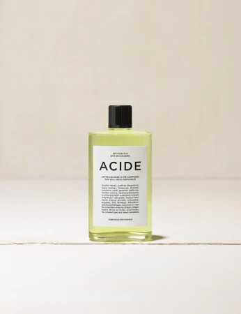 Close-up studio shot of a bottle of ACIDE eau de cologne on a white surface with a neutral-toned background.
