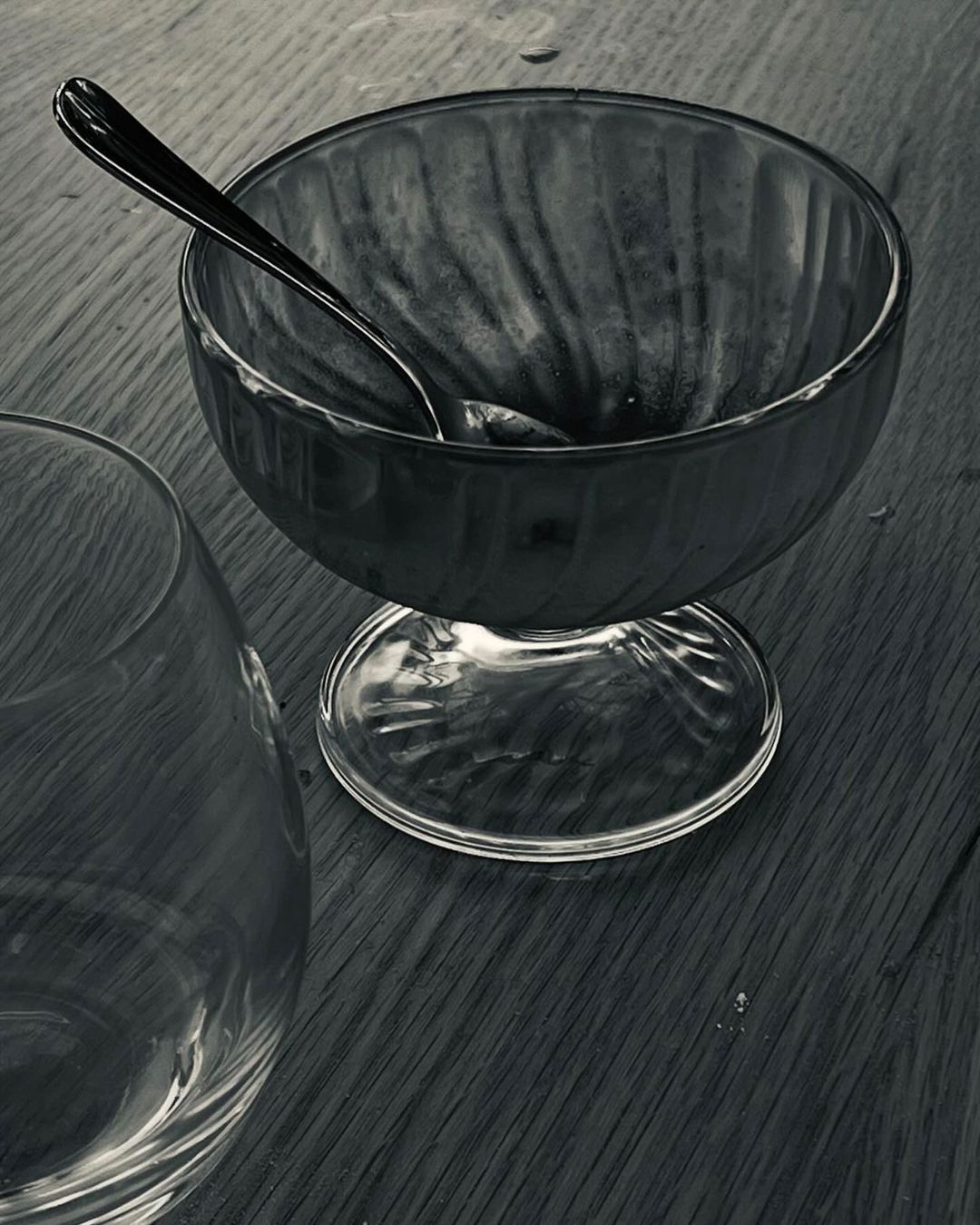 Close-up of a clear glass dessert bowl with a metal spoon resting inside, placed on a dark wooden table next to a glass.