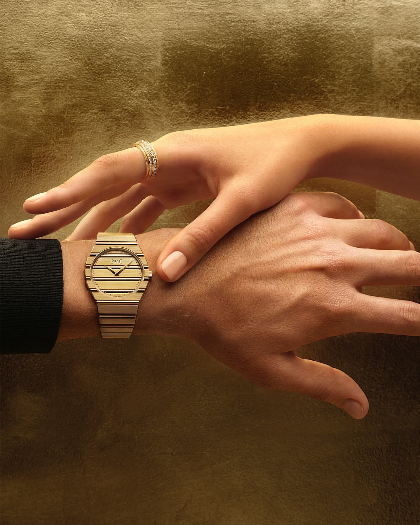 Close-up of two hands featuring a gold watch with a striped dial and a matching diamond ring against a golden textured background.