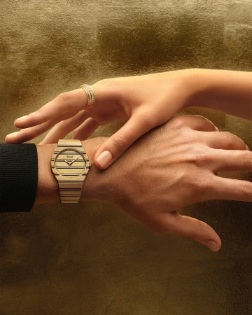 Close-up of two hands featuring a gold watch with a striped dial and a matching diamond ring against a golden textured background.