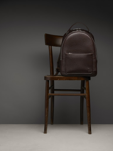 Dark brown leather backpack on a dark brown wooden chair against a gray background in a product studio shot for fashion.