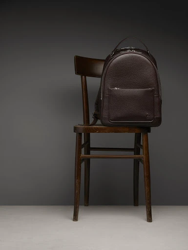 Dark brown leather backpack on a dark brown wooden chair against a gray background in a product studio shot for fashion.