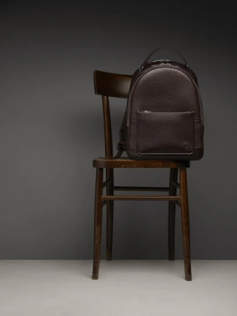 Dark brown leather backpack on a dark brown wooden chair against a gray background in a product studio shot for fashion.