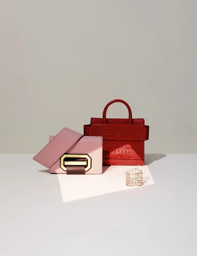 Still life studio shot of a pink handbag and a stacked red handbag with a gold metal accent piece on a white surface.