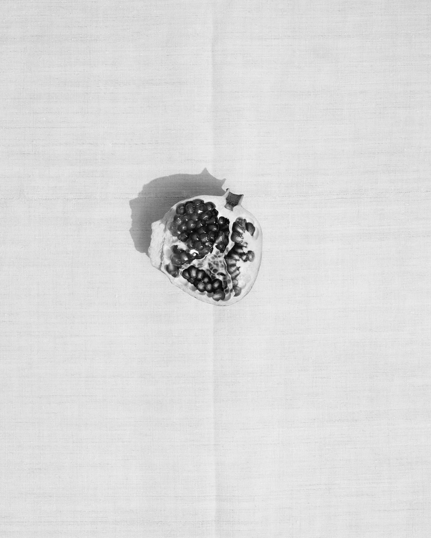 Monochrome close-up of a pomegranate half with visible seeds resting on a textured white surface with natural light casting a soft shadow.