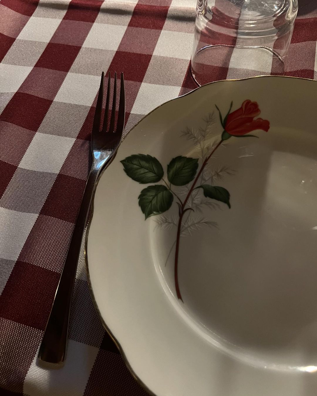 Overhead shot of a place setting with a white plate featuring a red rose design, a silver fork, and a clear glass on a red and white checkered tablecloth.