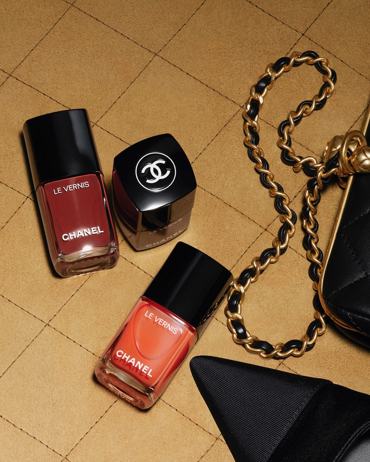 Chanel beauty products: nail polish in shades of coral and dark red arranged next to a handbag on a quilted surface.