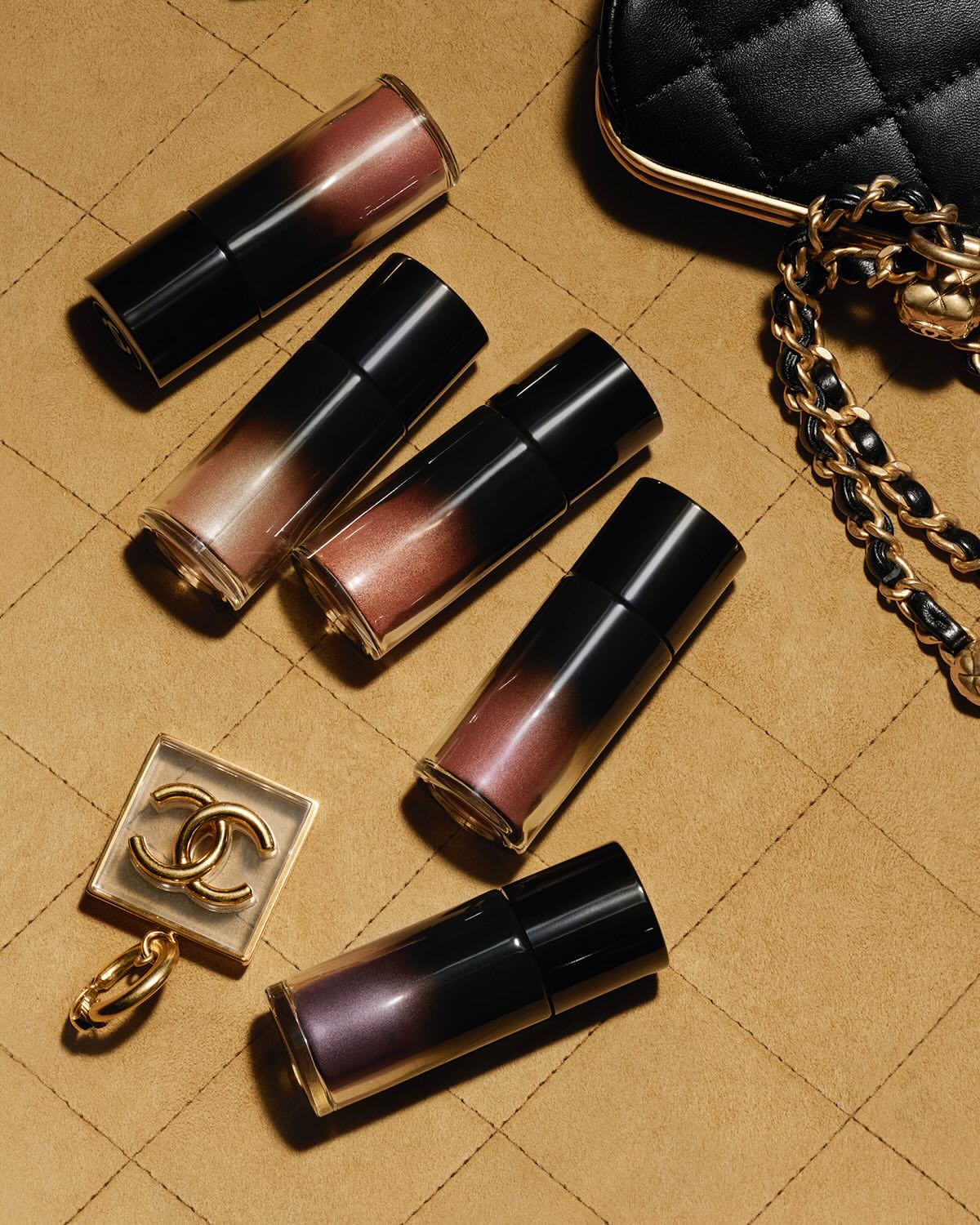 High-angle shot of Chanel lip gloss tubes with a quilted handbag and logo charm on a tan, quilted surface with good studio lighting.