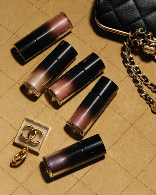High-angle shot of Chanel lip gloss tubes with a quilted handbag and logo charm on a tan, quilted surface with good studio lighting.