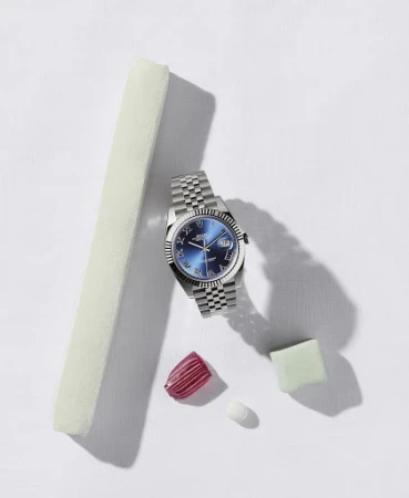 Wristwatch with a blue face and silver band displayed with decorative items including a pink nail and pale green cube on white.