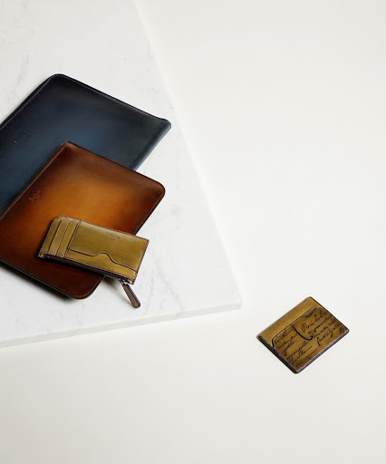 A flat lay captures leather goods: a blue document holder, a brown folder, and a smaller cardholder on white marble and surface.
