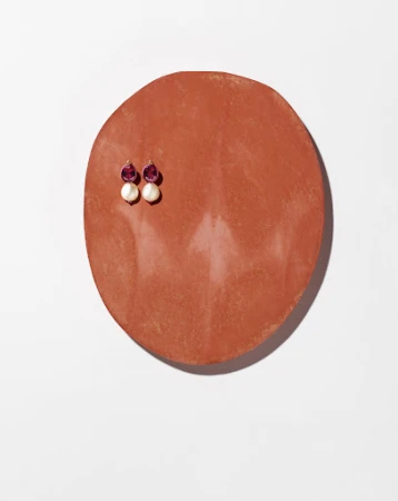 A pair of drop earrings with magenta stones and white pearls pinned to a terracotta-colored oval plaque on a white background.