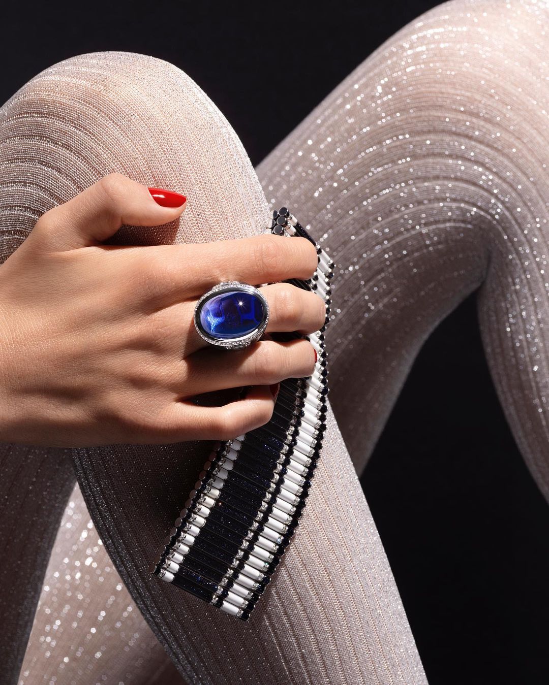 Close-up of a woman's hand wearing a blue gemstone ring, showcasing fashion jewelry with a black and white beaded clutch and shiny leggings.