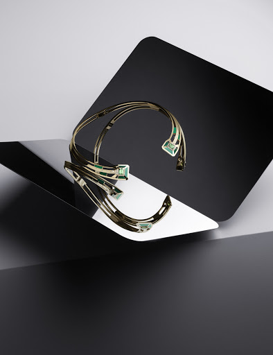 Elegant gold choker necklace with green gemstones displayed on a reflective surface with a geometric black and gray background in studio lighting.