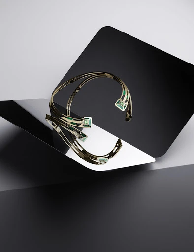Elegant gold choker necklace with green gemstones displayed on a reflective surface with a geometric black and gray background in studio lighting.