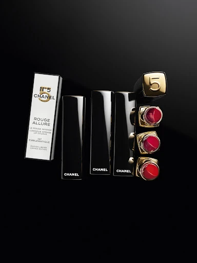 Elevated view of Chanel Rouge Allure lipstick tubes and box on a black surface. Gold and red lipstick shades.