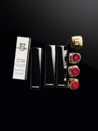 Elevated view of Chanel Rouge Allure lipstick tubes and box on a black surface. Gold and red lipstick shades.