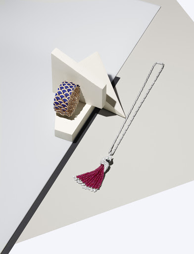 A blue and gold cuff bracelet sits on white geometric blocks next to a long silver tassel necklace on a grey and white surface.