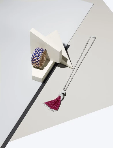 A blue and gold cuff bracelet sits on white geometric blocks next to a long silver tassel necklace on a grey and white surface.