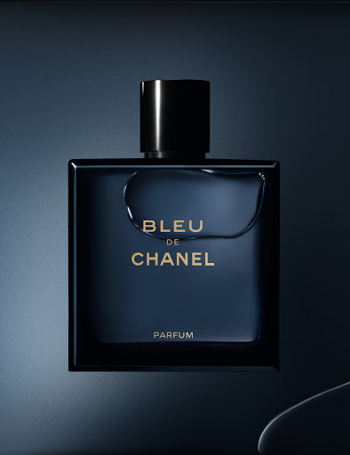 Bleu de Chanel Parfum bottle close-up. Black cap atop the square blue bottle, featuring silver lettering, stands against a shaded blue background.