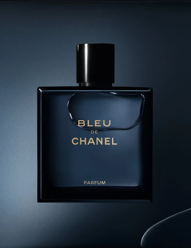 Bleu de Chanel Parfum bottle close-up. Black cap atop the square blue bottle, featuring silver lettering, stands against a shaded blue background.