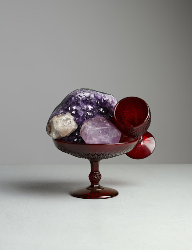 Still life of assorted amethyst crystals in a red pedestal bowl with matching goblets against a neutral grey background in a studio.