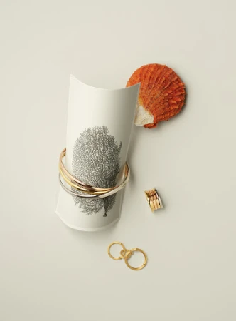 Elevated studio shot showcases a white decorative paper form with gold bangle bracelets, ring and keychain, accompanied by a seashell on a beige backdrop.