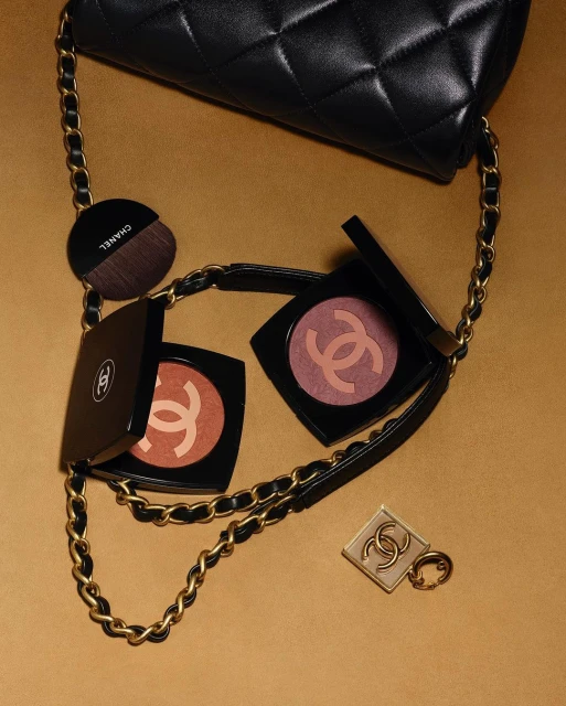 Luxury beauty still life featuring Chanel makeup compacts, blush brush, and a quilted leather handbag with a gold chain strap on gold background.