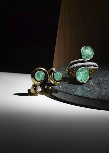 Elegant gold cuff bracelet and stud earrings featuring carved green gemstones displayed on a dark marble surface against a warm wood background.