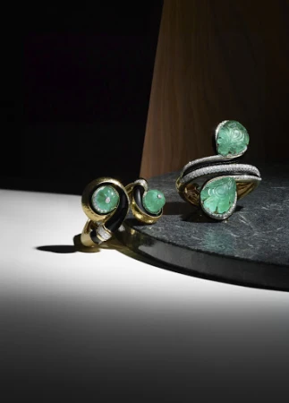 Elegant gold cuff bracelet and stud earrings featuring carved green gemstones displayed on a dark marble surface against a warm wood background.