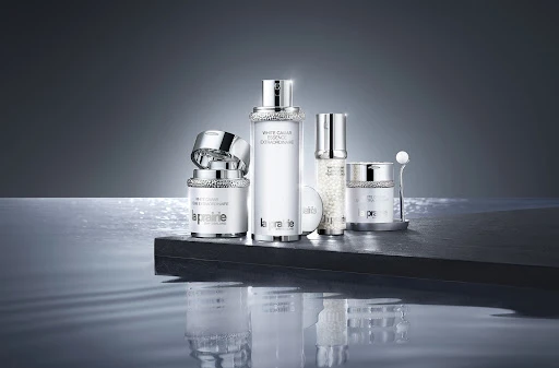 Luxury La Prairie skincare products in a studio shot feature white jars and bottles on a black surface with water reflections.