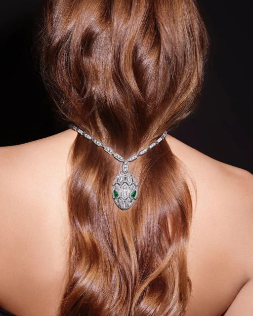 Woman's wavy brown hair cascading down her back with a Bulgari Serpenti necklace featuring diamonds and emeralds against a black background.