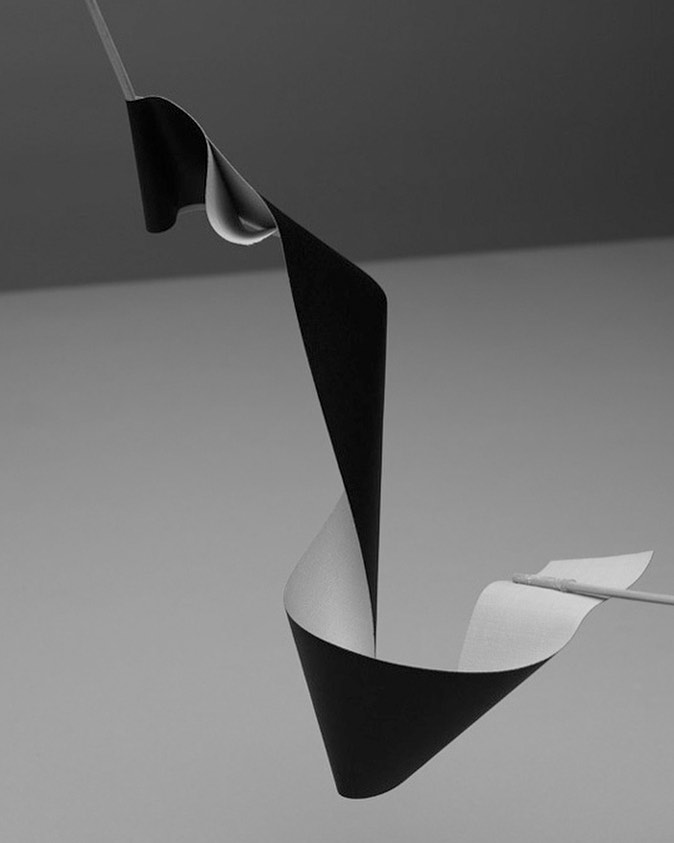 Minimalist studio shot of a curled black and white paper strip suspended by wooden rods against a gray backdrop, showcasing abstract design.