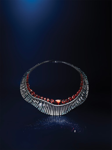Close-up of a silver necklace featuring red and clear gemstones against a blurred blue and black background with light reflections.