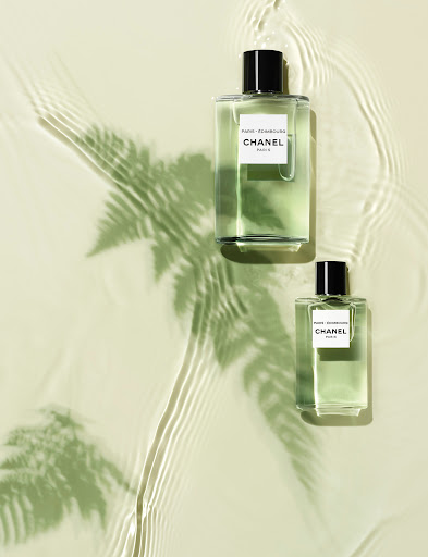 Two clear glass Chanel perfume bottles with black caps sit on a pale green surface with a fern shadow and water ripple patterns.