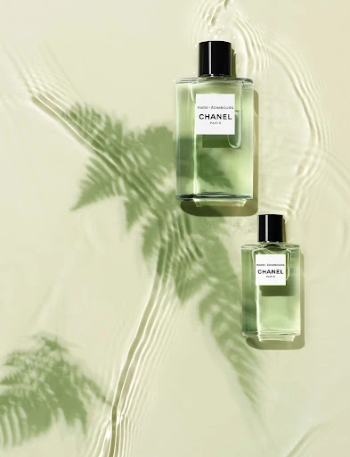 Two clear glass Chanel perfume bottles with black caps sit on a pale green surface with a fern shadow and water ripple patterns.