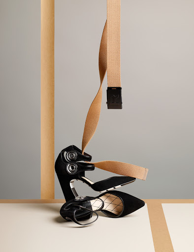 Black high heel shoe sits on a beige surface with a tan belt suspended above it, set against a neutral gray background.