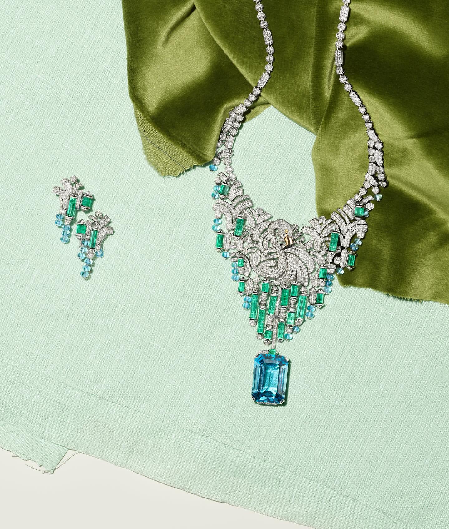 Elegant jewelry featuring a diamond necklace with blue and green gemstones alongside matching earrings, set against a pastel green fabric backdrop.