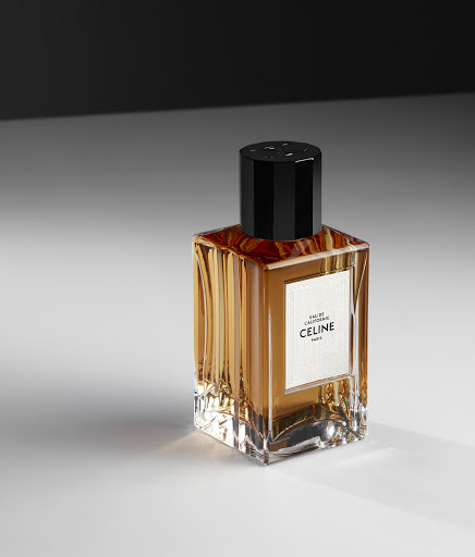 Celine perfume bottle with rectangular glass body and black cap on a white surface, against a dark blurred background.